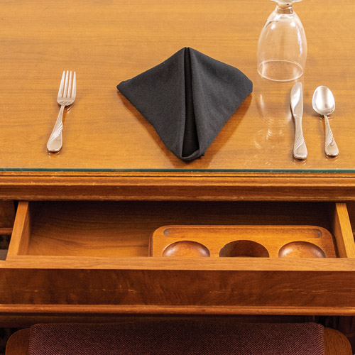 table with a glass, napkin, and silverware on it along with a open drawer with a tray in it