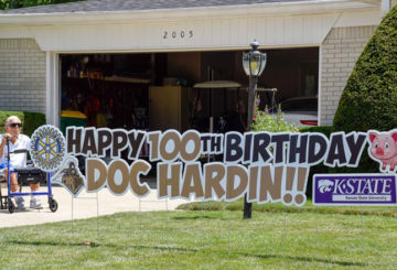 Doc Hardin sits in his driveway next to a large sign that says Happy 100th Birthday Doc Hardin with a Rotary Club symbol, Purdue Boilermaker Special logo, and Kansas State sign. There is also a pig to represent his farm and veterinary career.