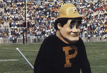 1980s era Purdue Pete in a Block P sweatshirt and hardhat walking across the field at Ross-Ade Stadium with a crowd of fans in the bleachers behind him.