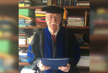 James Franklin Sharp stands in his PhD cap and gown in front of a bookshelf in his NYC apartment